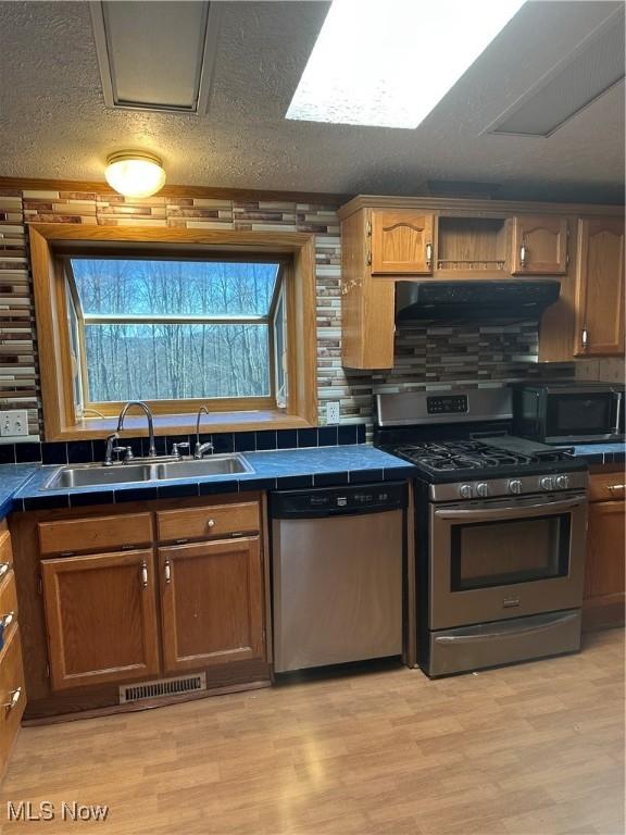 kitchen with sink, appliances with stainless steel finishes, tile counters, light hardwood / wood-style floors, and exhaust hood