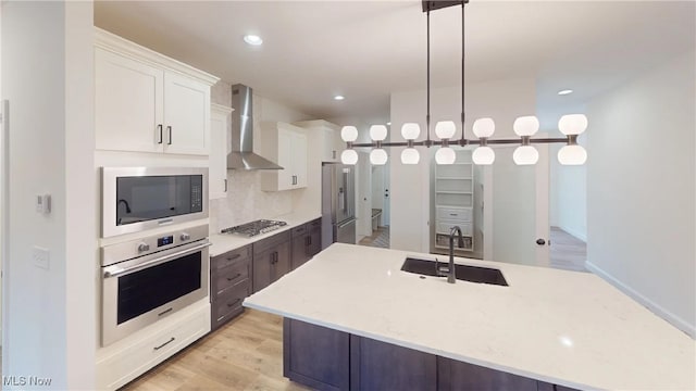 kitchen with hanging light fixtures, stainless steel appliances, sink, white cabinets, and wall chimney range hood