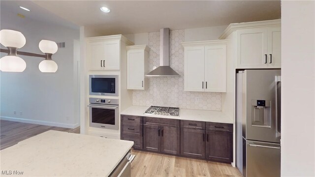 kitchen with appliances with stainless steel finishes, wall chimney exhaust hood, dark brown cabinetry, decorative light fixtures, and backsplash