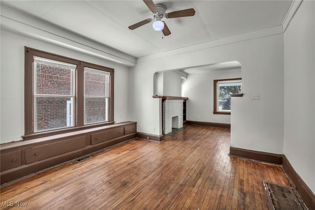 unfurnished room featuring arched walkways, ceiling fan, wood finished floors, baseboards, and a brick fireplace