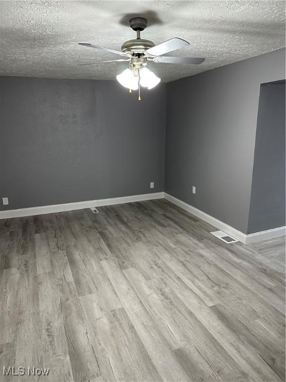 empty room with a textured ceiling, a ceiling fan, visible vents, baseboards, and light wood-type flooring