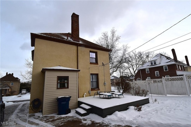 snow covered rear of property with a chimney, fence, and stucco siding