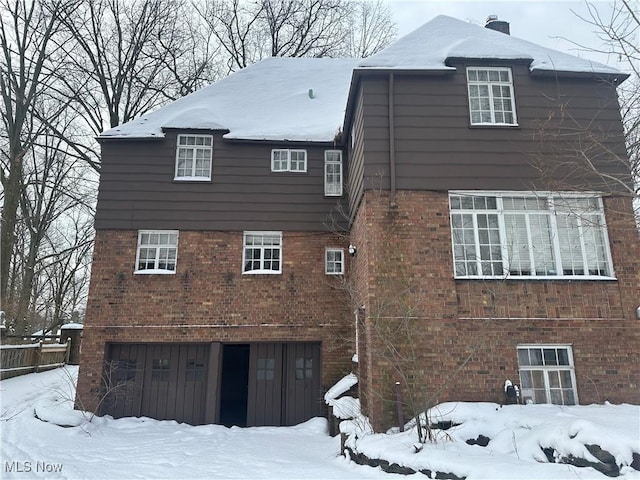 snow covered house with a garage, brick siding, and a chimney