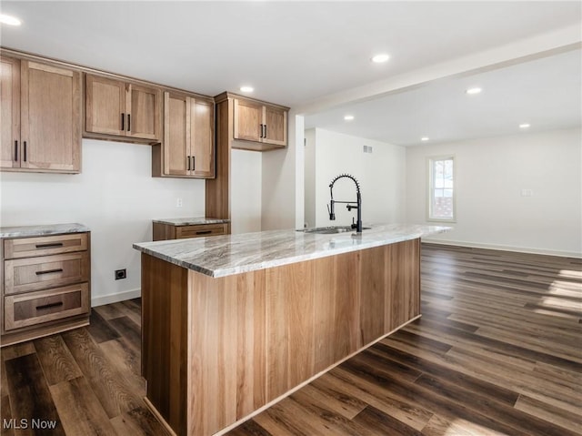 kitchen with light stone counters, a sink, brown cabinets, dark wood-style floors, and a center island with sink