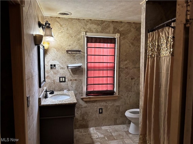 bathroom featuring stone tile floors, visible vents, toilet, vanity, and tile walls