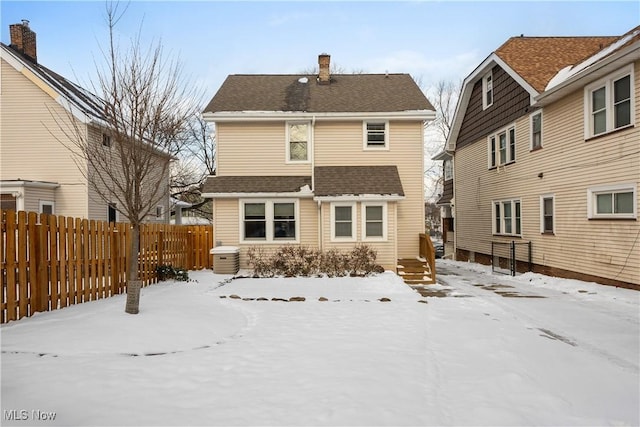 snow covered house with a fenced backyard, a chimney, and roof with shingles