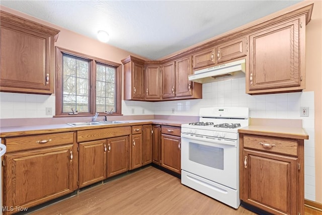 kitchen featuring white range with gas stovetop, under cabinet range hood, a sink, light countertops, and brown cabinetry