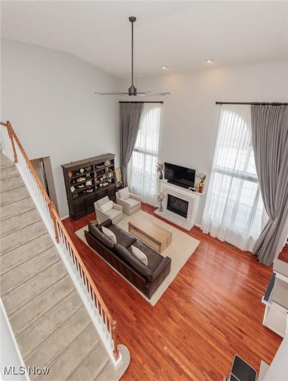 living room featuring stairway, a glass covered fireplace, ceiling fan, vaulted ceiling, and wood finished floors