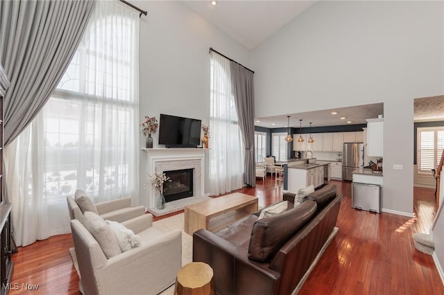 living room with high vaulted ceiling, a glass covered fireplace, baseboards, and wood finished floors