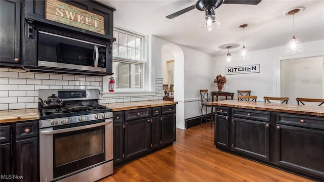 kitchen featuring arched walkways, wood finished floors, decorative light fixtures, stainless steel appliances, and dark cabinetry