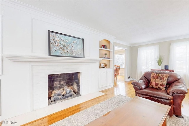 living area with light wood-type flooring, a fireplace with flush hearth, built in shelves, and crown molding