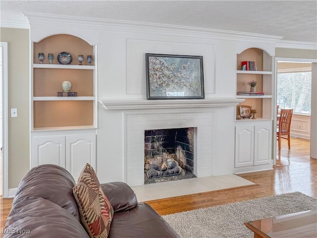 living room with a textured ceiling, built in shelves, a fireplace, light wood-type flooring, and crown molding