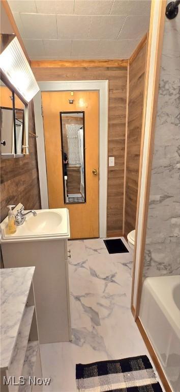 bathroom with a bathtub, marble finish floor, toilet, wood walls, and vanity