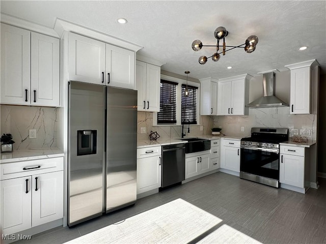 kitchen featuring a sink, white cabinets, wall chimney range hood, appliances with stainless steel finishes, and pendant lighting