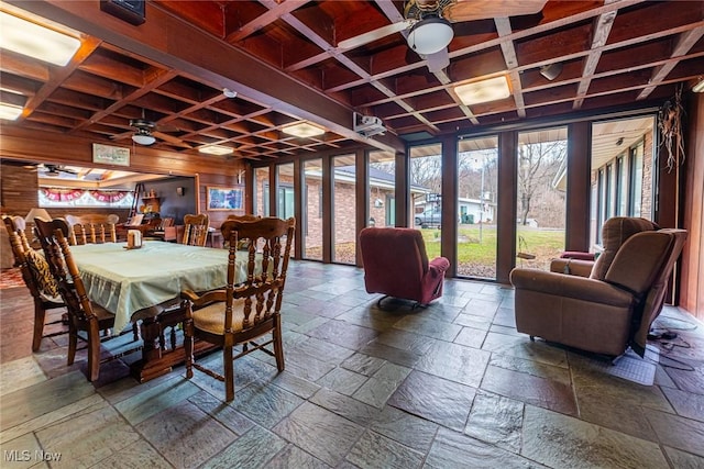 dining area featuring wood walls, stone tile floors, coffered ceiling, and a ceiling fan