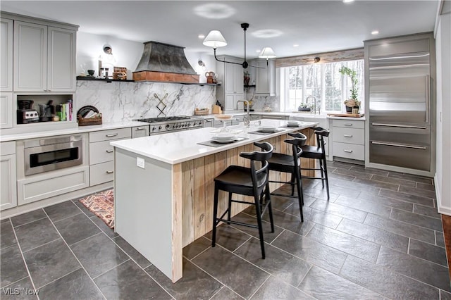 kitchen featuring a breakfast bar, a center island with sink, stainless steel appliances, light countertops, and custom range hood