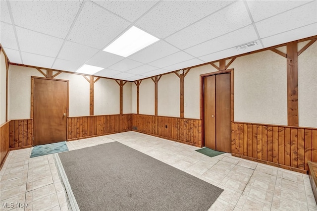 unfurnished room featuring light floors, visible vents, wainscoting, wooden walls, and a drop ceiling