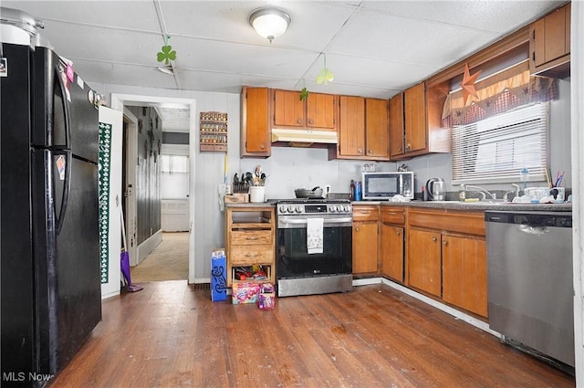 kitchen with appliances with stainless steel finishes, brown cabinets, dark wood-style flooring, under cabinet range hood, and a sink