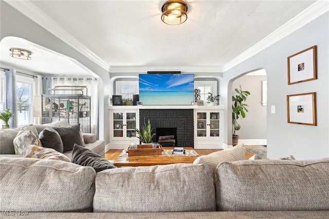 living room featuring baseboards, arched walkways, ornamental molding, wood finished floors, and a brick fireplace