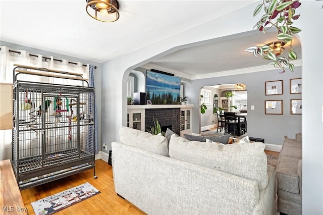 living room featuring baseboards, arched walkways, ornamental molding, wood finished floors, and a brick fireplace