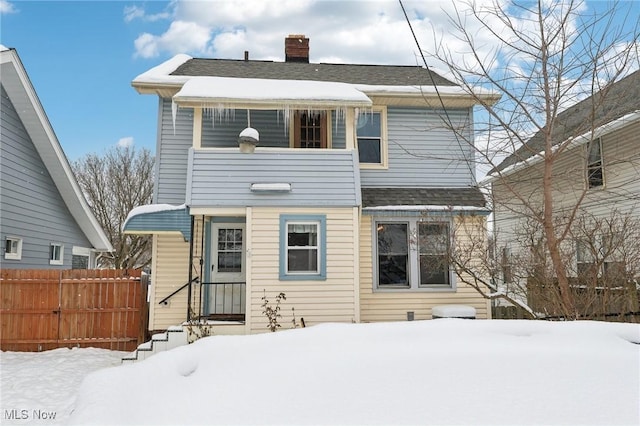 snow covered house featuring roof with shingles, fence, and a chimney