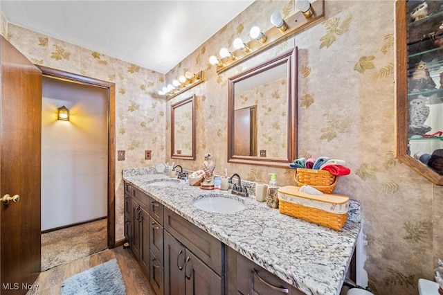 bathroom with double vanity, wallpapered walls, a sink, and wood finished floors