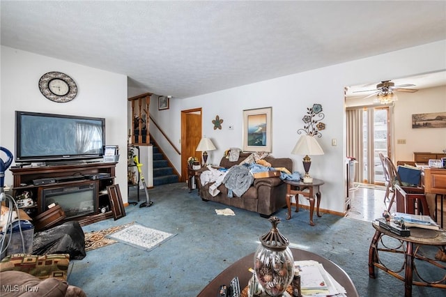 carpeted living room with ceiling fan, a textured ceiling, baseboards, stairs, and a glass covered fireplace