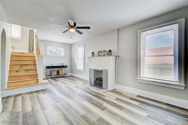 unfurnished living room featuring a ceiling fan, baseboards, stairway, a brick fireplace, and light wood finished floors