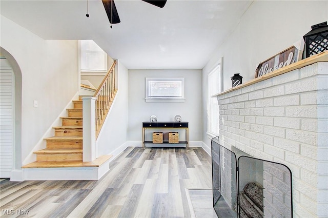 living room featuring a ceiling fan, baseboards, stairs, a brick fireplace, and light wood-type flooring