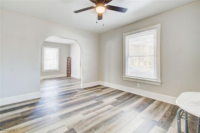 empty room featuring arched walkways, a ceiling fan, light wood-style flooring, and baseboards