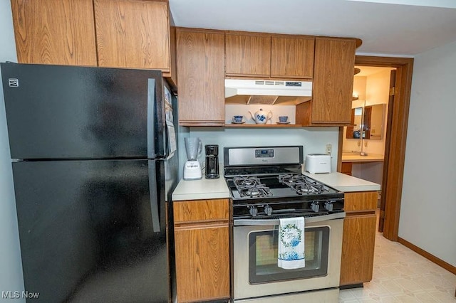 kitchen with baseboards, freestanding refrigerator, light countertops, under cabinet range hood, and stainless steel range with gas stovetop