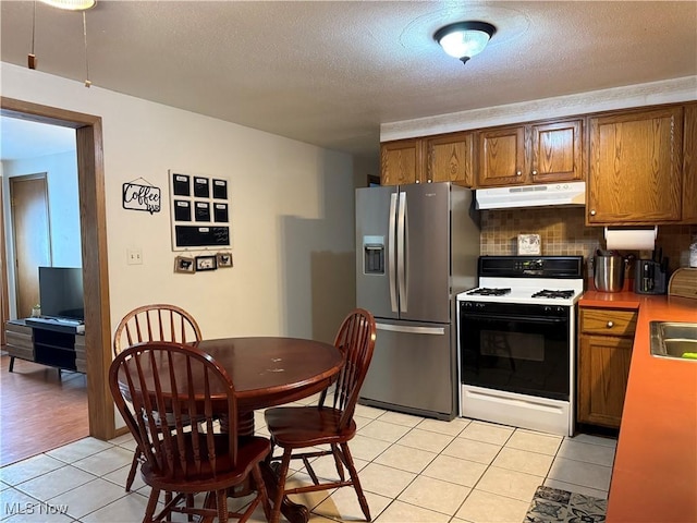 kitchen featuring range with gas stovetop, brown cabinetry, light tile patterned flooring, stainless steel fridge, and under cabinet range hood