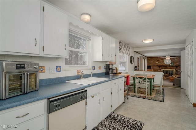 kitchen featuring white dishwasher, a sink, white cabinets, a brick fireplace, and light floors