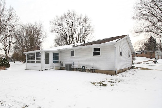 snow covered rear of property with a sunroom, a chimney, and central AC unit