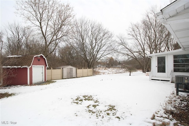 yard layered in snow featuring a storage shed, central AC, fence, a garage, and an outdoor structure