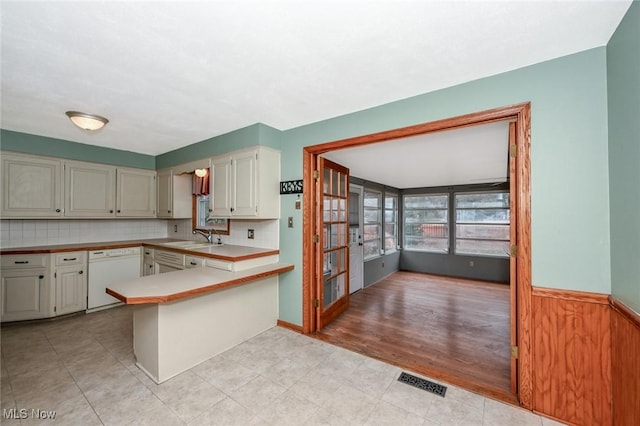 kitchen with white dishwasher, a peninsula, a sink, light countertops, and wainscoting