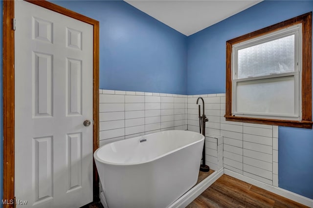 full bathroom featuring a soaking tub, a wainscoted wall, tile walls, and wood finished floors