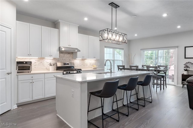 kitchen with under cabinet range hood, stove, a sink, light countertops, and a center island with sink