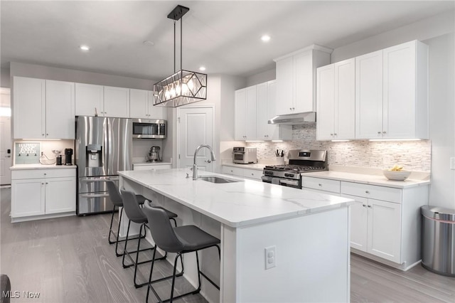 kitchen with a center island with sink, stainless steel appliances, white cabinetry, a sink, and under cabinet range hood