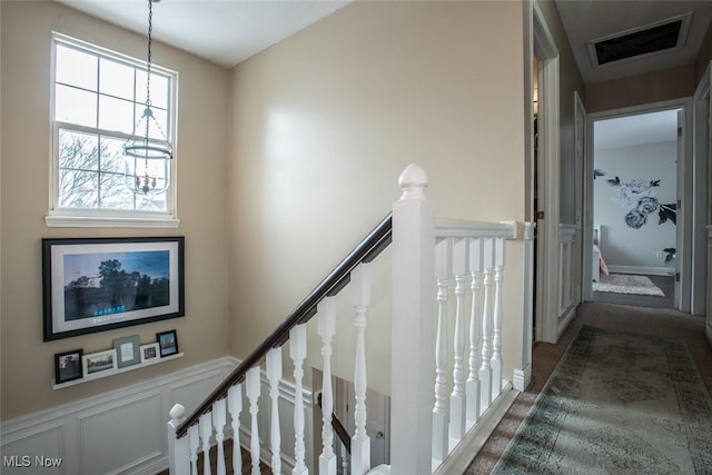 hallway with a wainscoted wall, a decorative wall, visible vents, and an upstairs landing