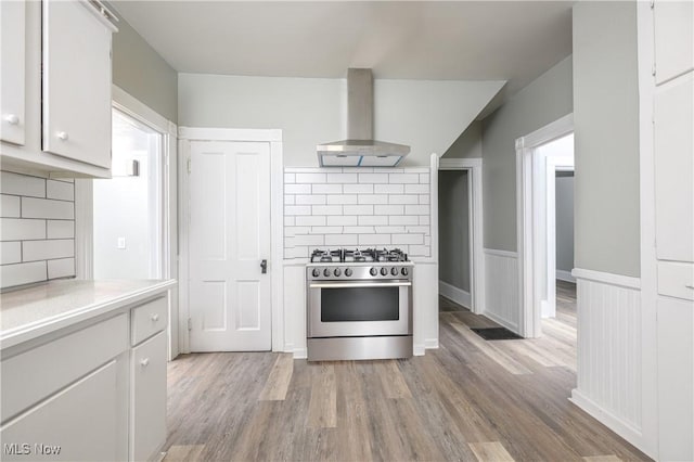 kitchen featuring white cabinets, light countertops, wainscoting, stainless steel range with gas cooktop, and wall chimney exhaust hood