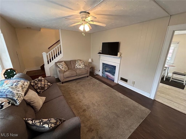 living area with stairs, a ceiling fan, a fireplace with raised hearth, and dark wood-style flooring