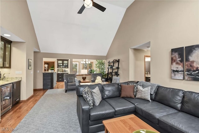 living room with indoor wet bar, light wood finished floors, high vaulted ceiling, beverage cooler, and ceiling fan with notable chandelier