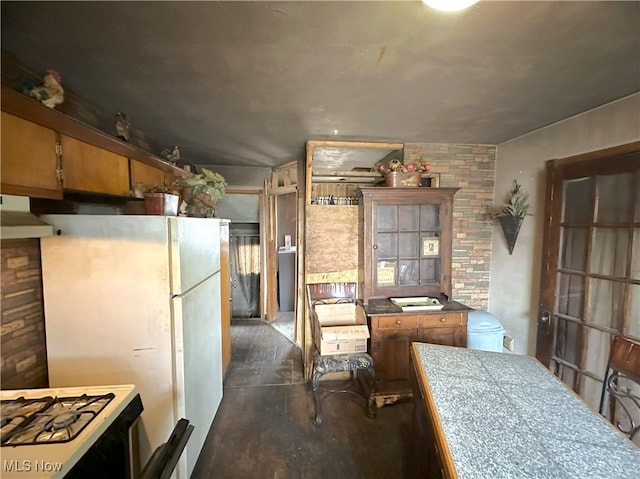 kitchen featuring under cabinet range hood, concrete floors, freestanding refrigerator, tile counters, and brown cabinetry