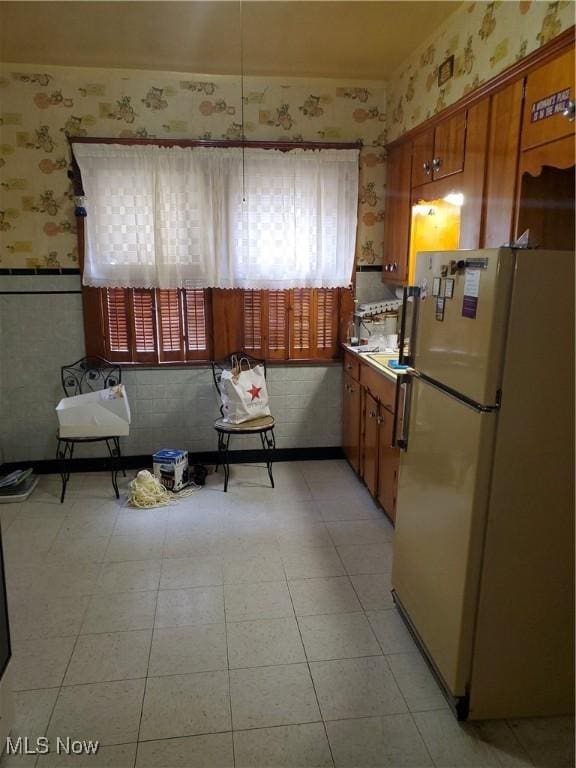 kitchen with wallpapered walls, wainscoting, brown cabinets, freestanding refrigerator, and light tile patterned flooring