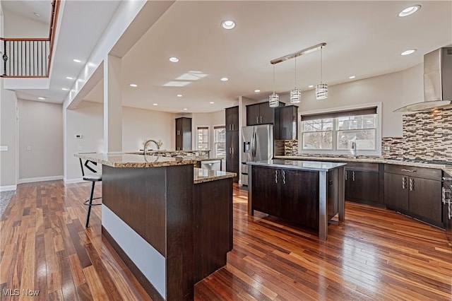 kitchen with stainless steel appliances, wall chimney range hood, a center island with sink, and dark brown cabinetry