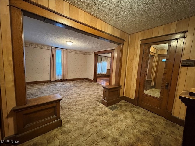 carpeted spare room featuring a textured ceiling, wooden walls, baseboards, and wallpapered walls