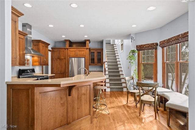 kitchen featuring light countertops, brown cabinetry, freestanding refrigerator, gas range, and wall chimney exhaust hood