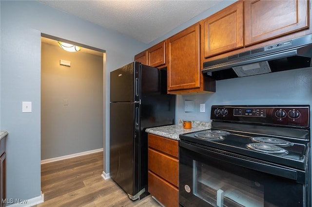 kitchen with brown cabinets, light wood finished floors, a textured ceiling, under cabinet range hood, and black appliances