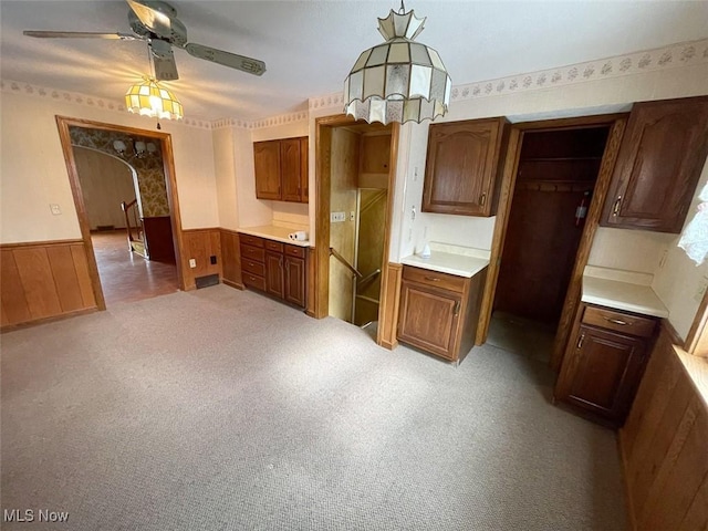 kitchen with arched walkways, a wainscoted wall, light countertops, light carpet, and ceiling fan